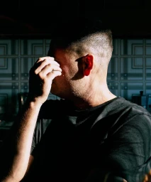 Man wearing a black t-shirt covering his face with his hand under strong directional lighting.