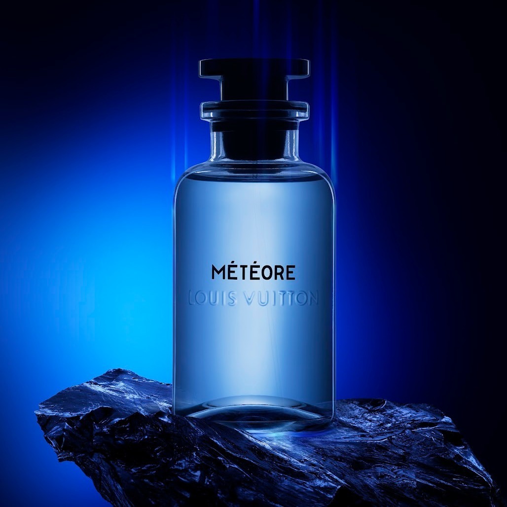 Close-up studio shot of a clear glass Louis Vuitton Météore perfume bottle on a black stone, illuminated by blue and black studio lighting.
