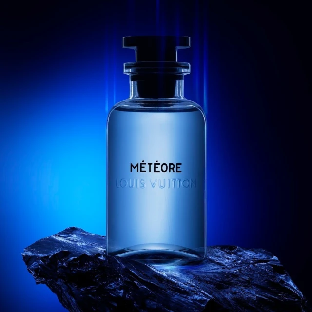 Close-up studio shot of a clear glass Louis Vuitton Météore perfume bottle on a black stone, illuminated by blue and black studio lighting.