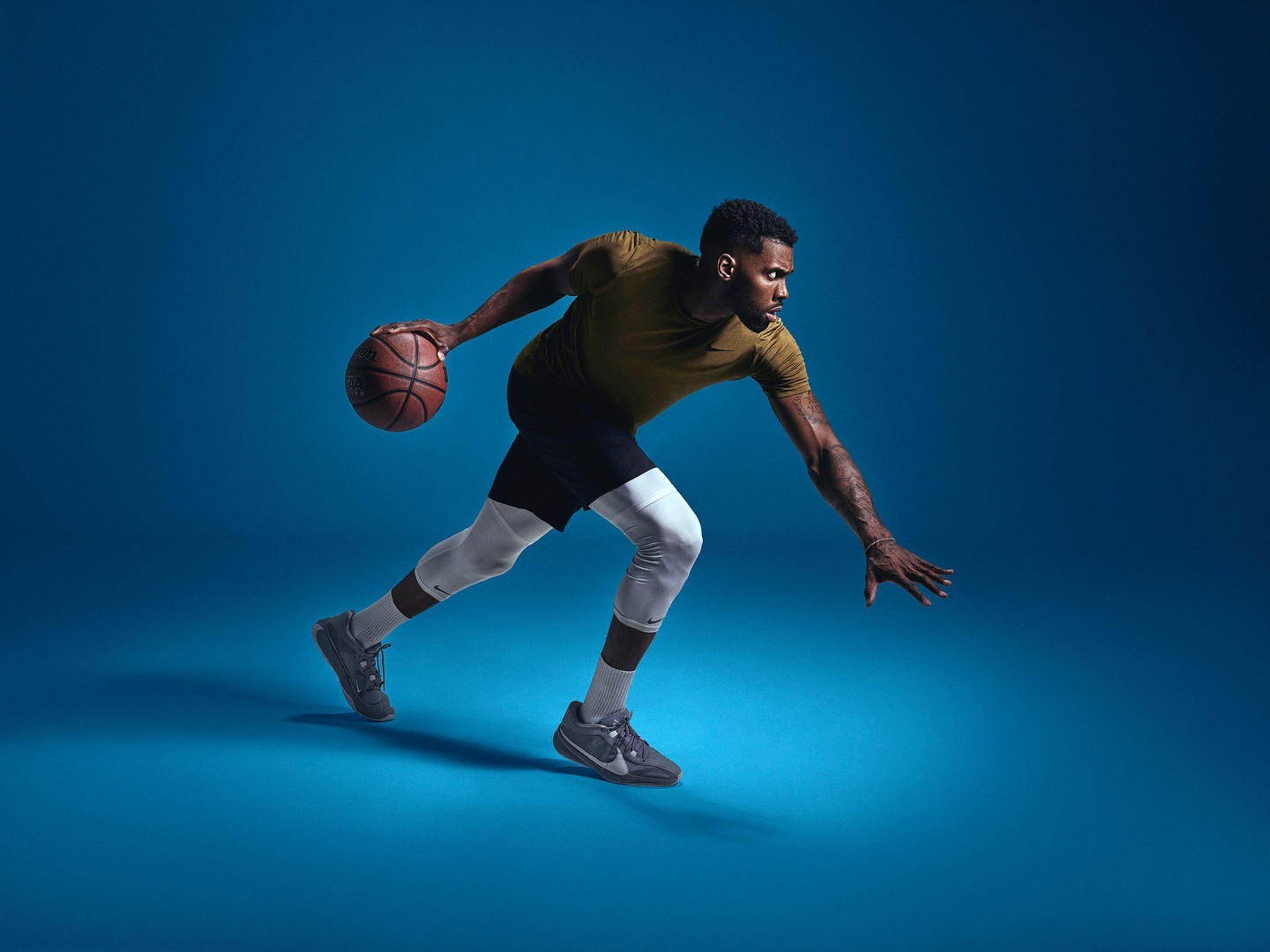 A male basketball player wearing a sports shirt, black shorts, compression leggings, and sneakers dribbles a basketball on blue backdrop.