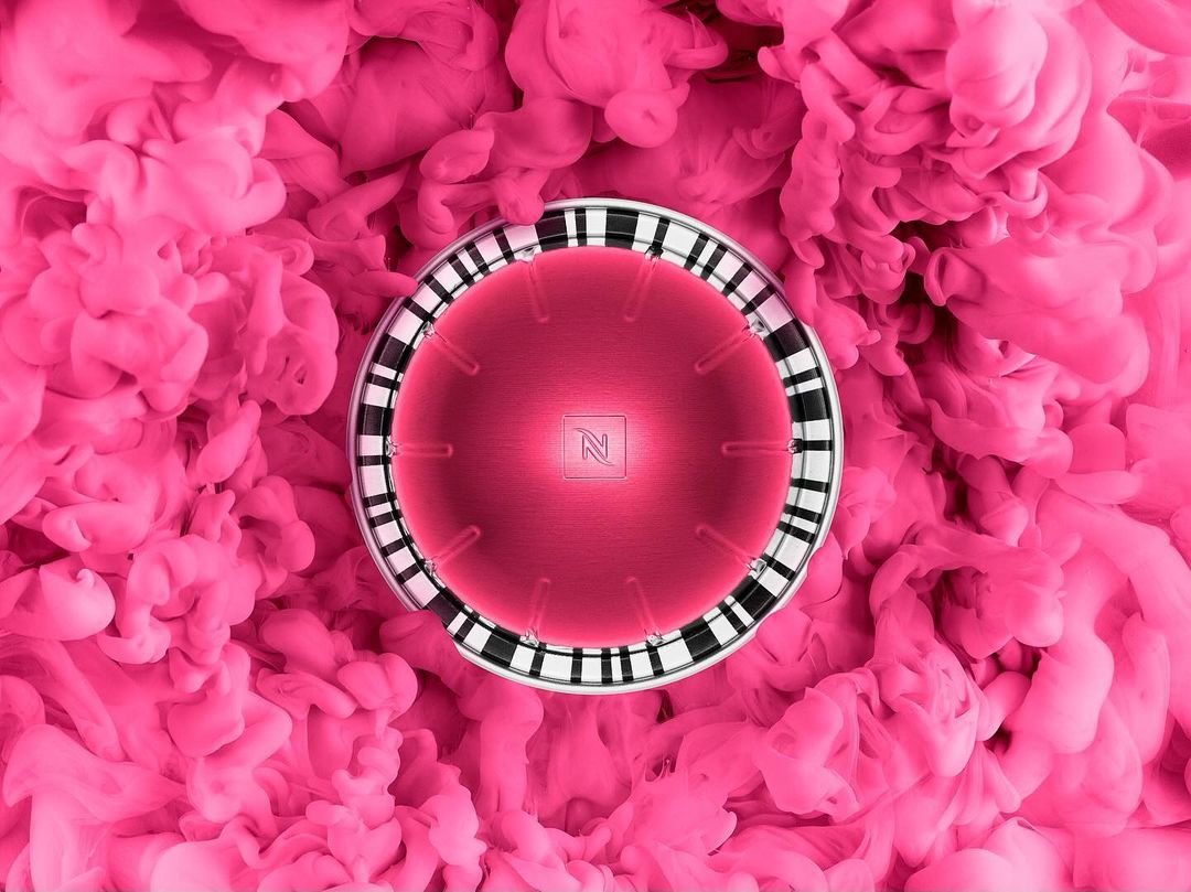 Pink coffee pod with Nespresso logo surrounded by pink smoke. Close-up studio shot emphasizing colors and textures.