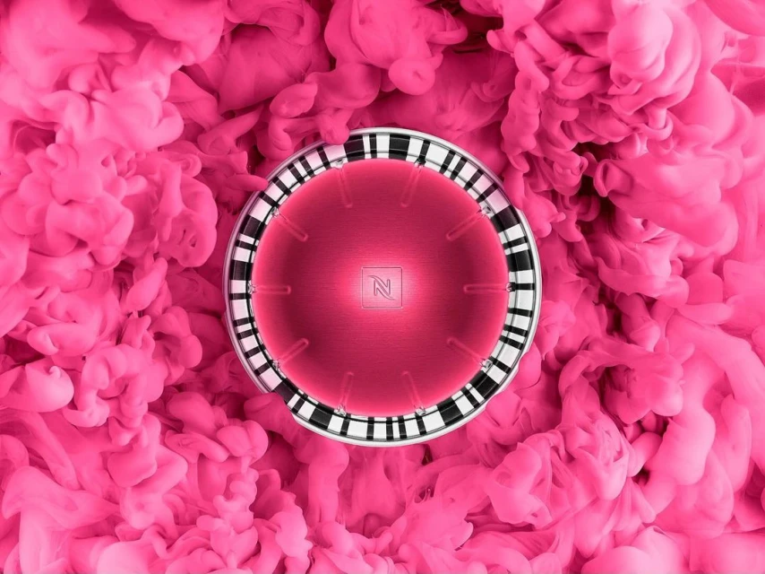 Pink coffee pod with Nespresso logo surrounded by pink smoke. Close-up studio shot emphasizing colors and textures.