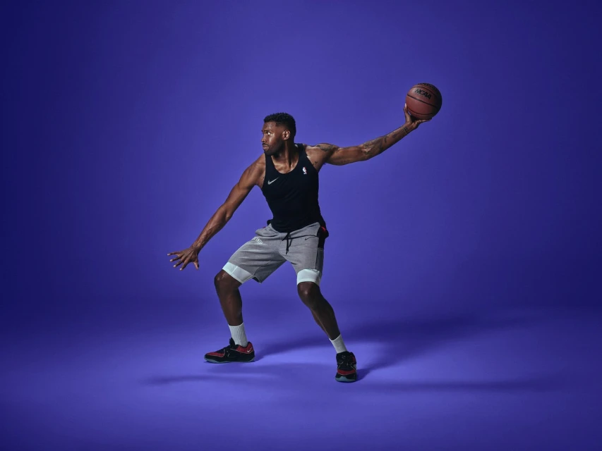 Male athlete in a tank top and shorts dribbles a basketball on a violet background in a studio setting.
