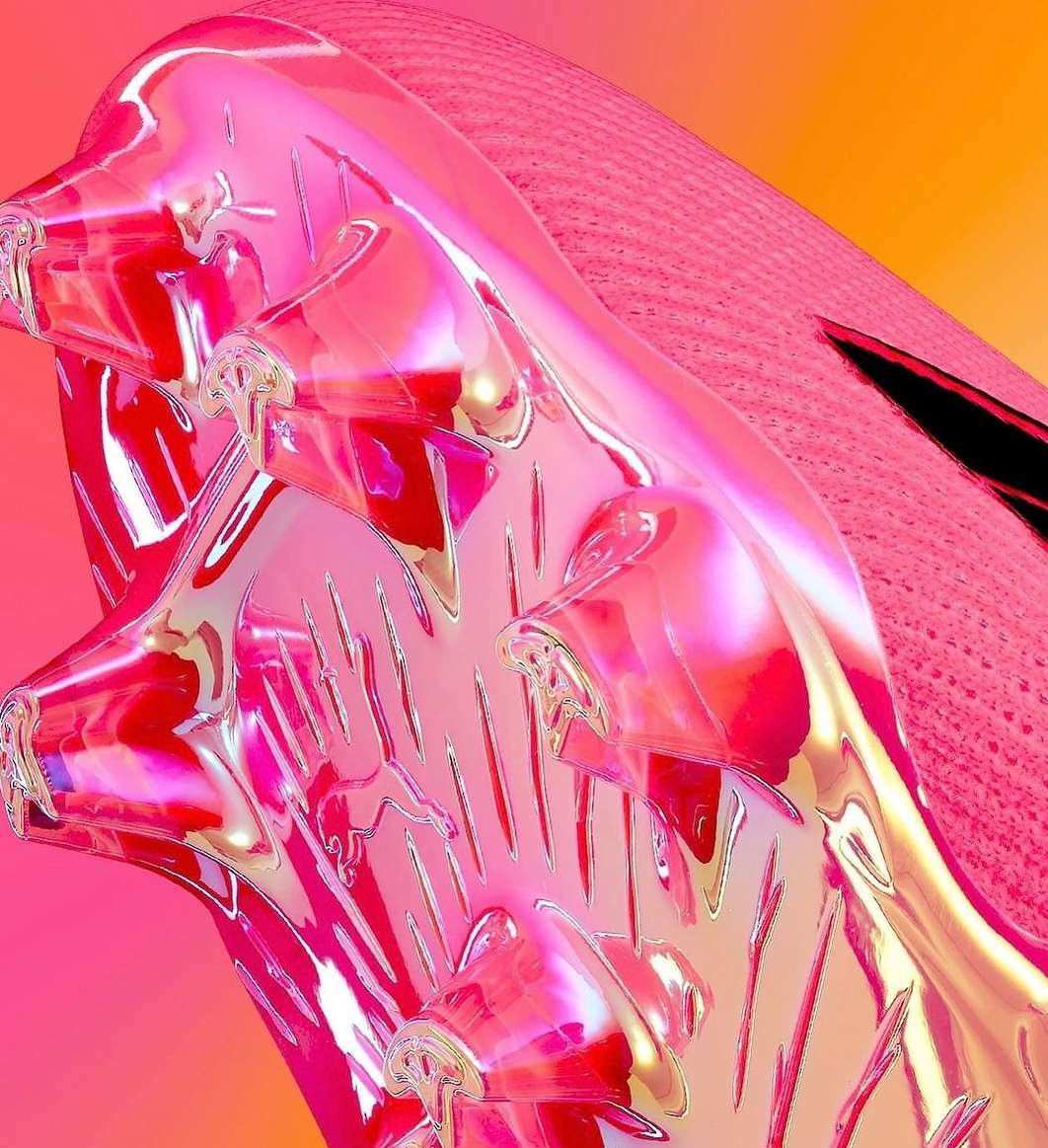 Close-up of a pink and iridescent soccer shoe cleat with pink knit upper, showing studs on a vibrant, blended orange and pink background.
