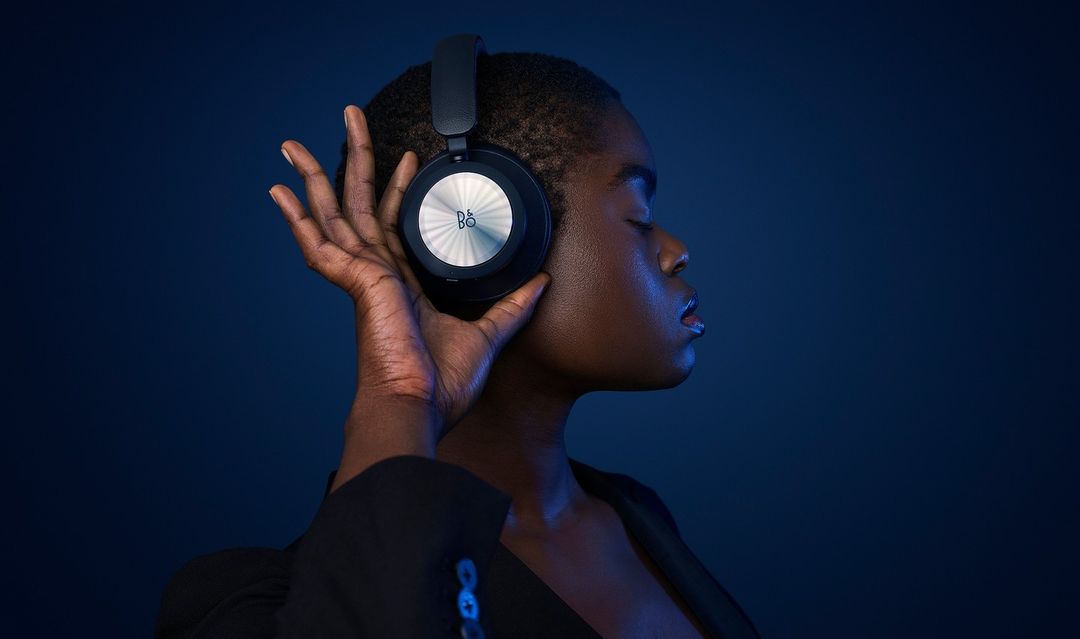 Woman with dark skin and short hair wearing headphones with a silver logo, hand near ear, dark blue background, low-key lighting.
