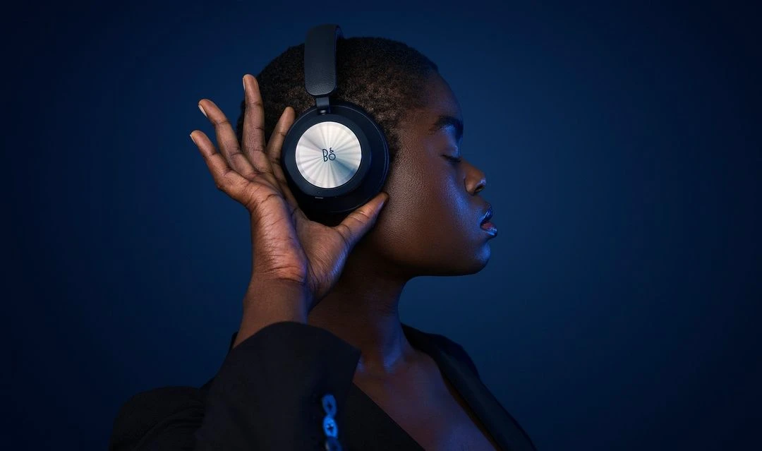 Woman with dark skin and short hair wearing headphones with a silver logo, hand near ear, dark blue background, low-key lighting.