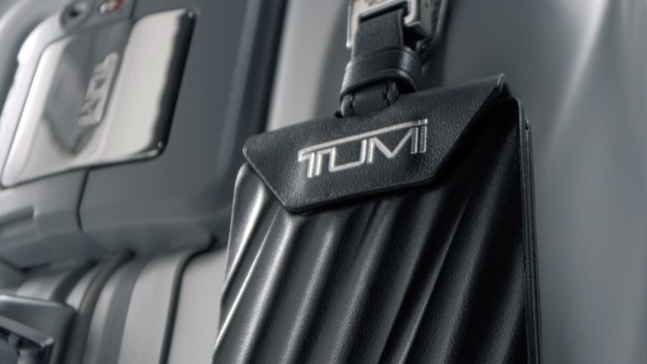 Close-up of a black leather TUMI luggage tag attached to a gray suitcase in an indoor setting with muted lighting.
