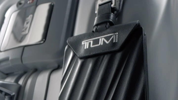 Close-up of a black leather TUMI luggage tag attached to a gray suitcase in an indoor setting with muted lighting.