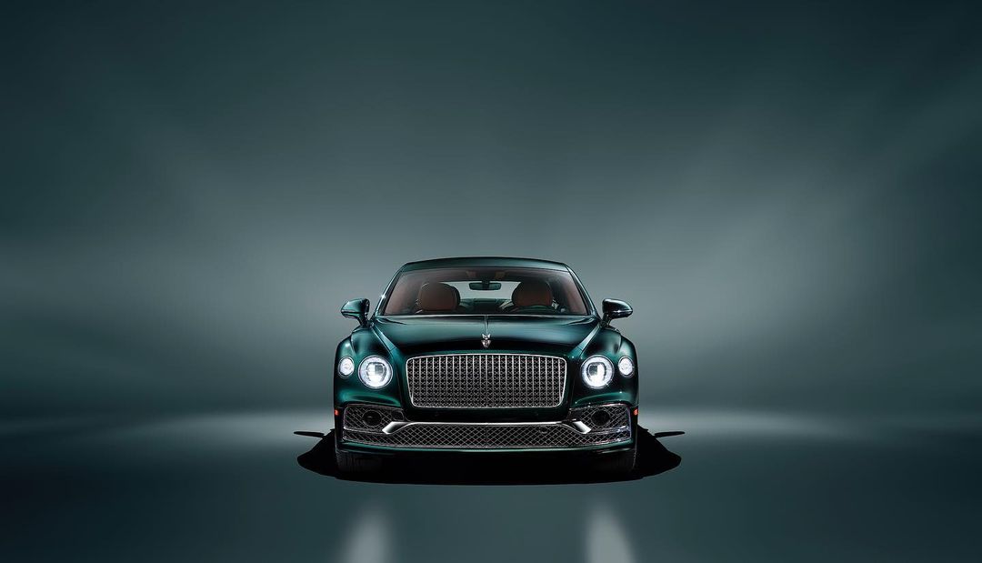 Front view of a dark green Bentley Flying Spur sedan against a blurred gray background in a studio setting with soft lighting.