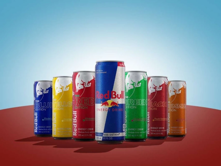 Assorted Red Bull energy drink cans in various flavors, including Blue Edition, Yellow Edition, Red Edition, Green Edition, and Peach Edition, on a red surface.