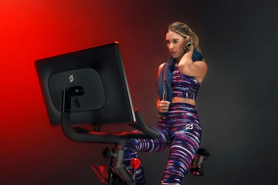 Adult female wearing fitness apparel using an exercise bike in a studio with a red background. She has a towel around her neck.