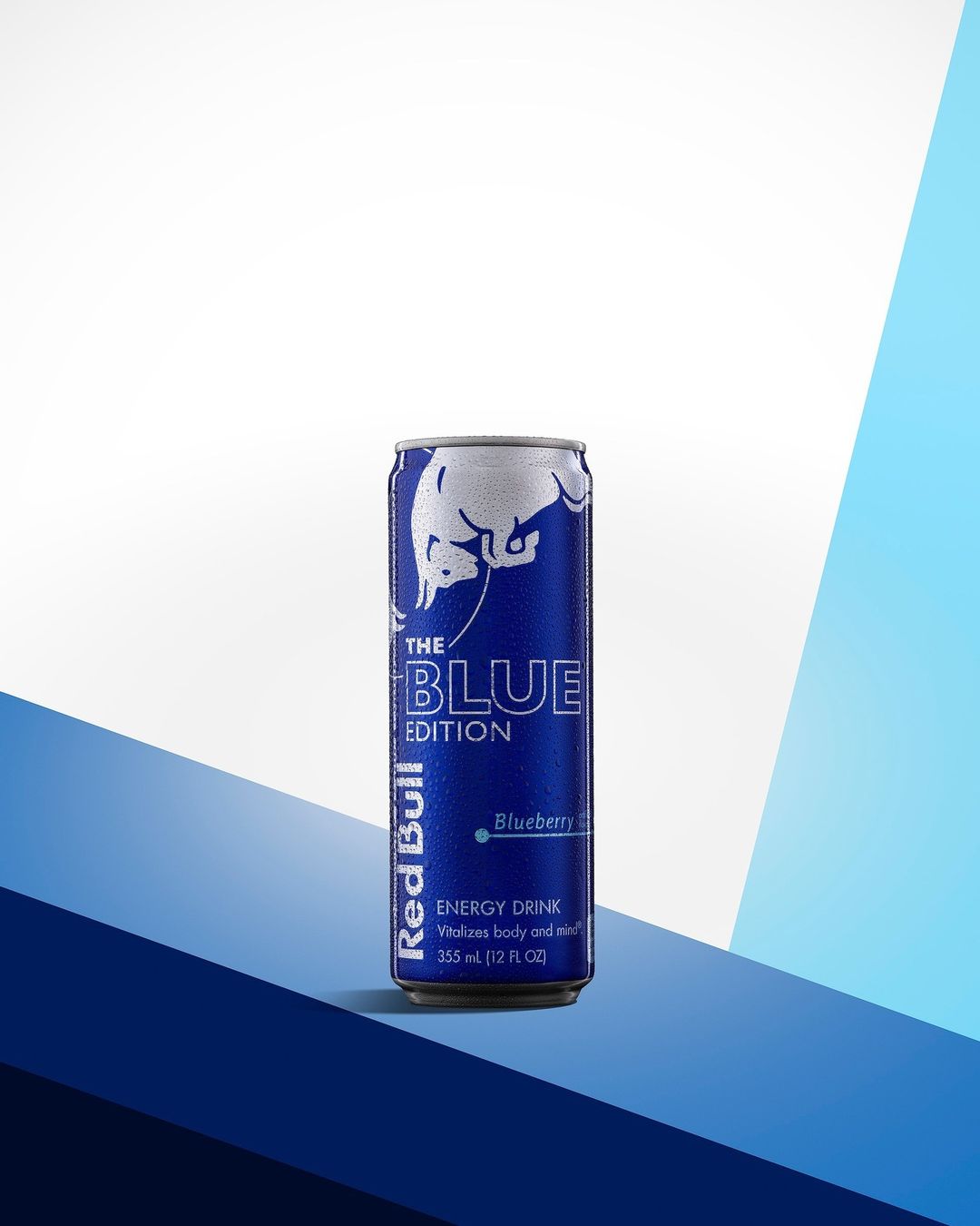 A single can of Red Bull The Blue Edition Blueberry energy drink on a blue and white geometric surface. Clean product shot.