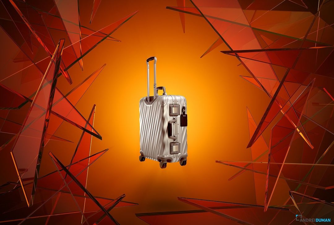 Studio shot of a silver suitcase with a retractable handle, surrounded by red-orange geometric shards on an orange gradient background.
