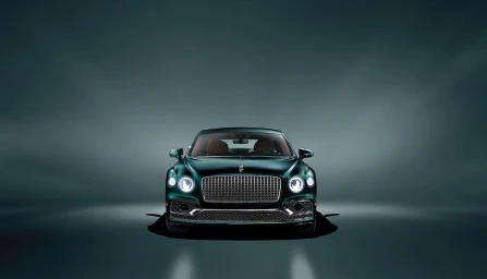 Front view of a dark green luxury car with illuminated headlights and a prominent chrome grille against a moody dark background.