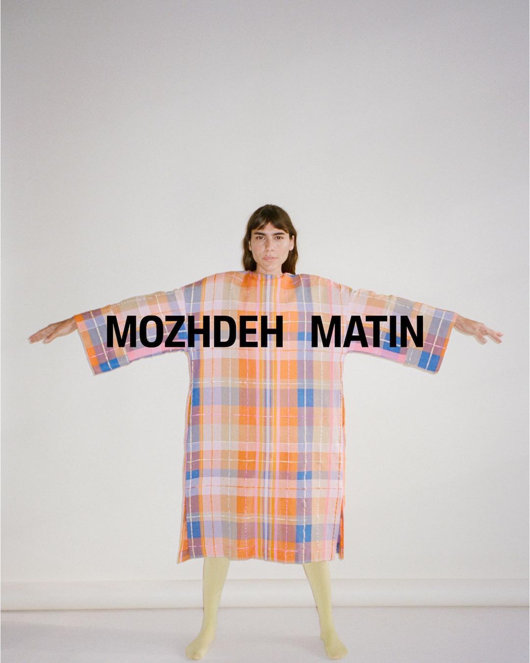 An adult woman with outstretched arms stands in front of a white background, wearing a colorful plaid dress with short yellow tights.