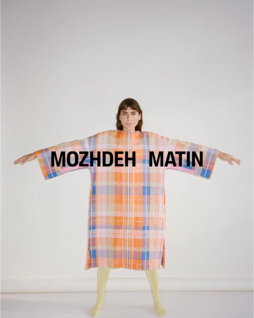 An adult woman with outstretched arms stands in front of a white background, wearing a colorful plaid dress with short yellow tights.