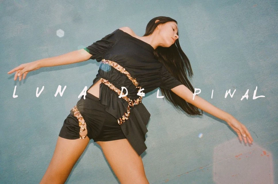 Woman in a black outfit posing in front of a textured teal wall, arms outstretched, with the text 'Luna Del Pinal' overlaid.