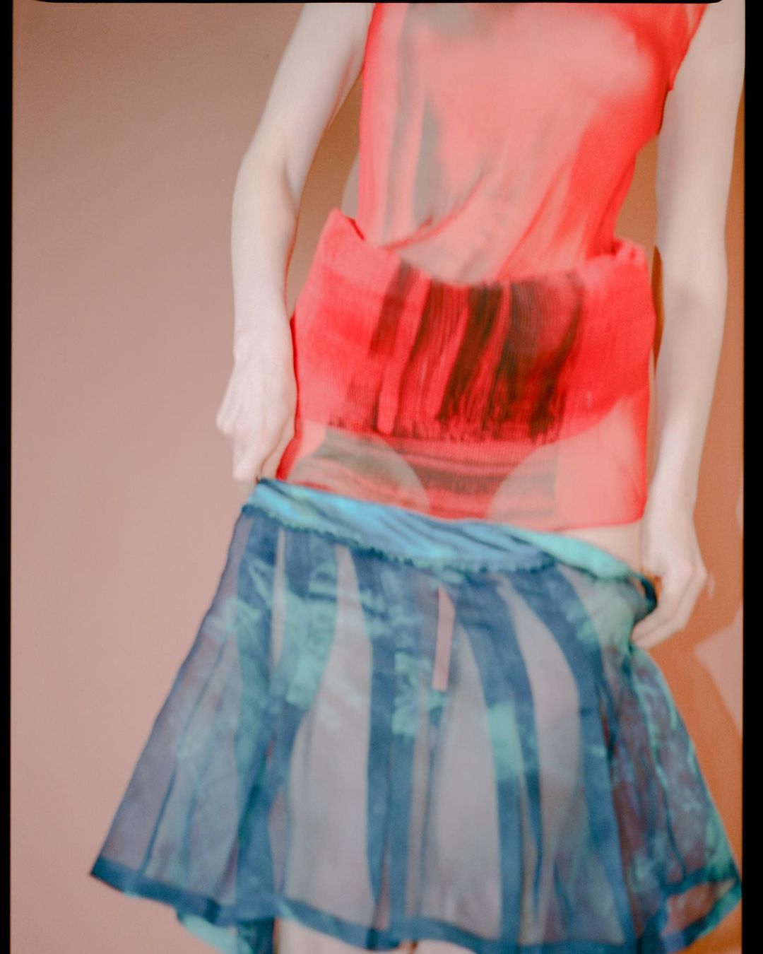 A woman in an orange top and blue skirt stands against a peach background, adjusting her skirt. The composition is bright and airy.