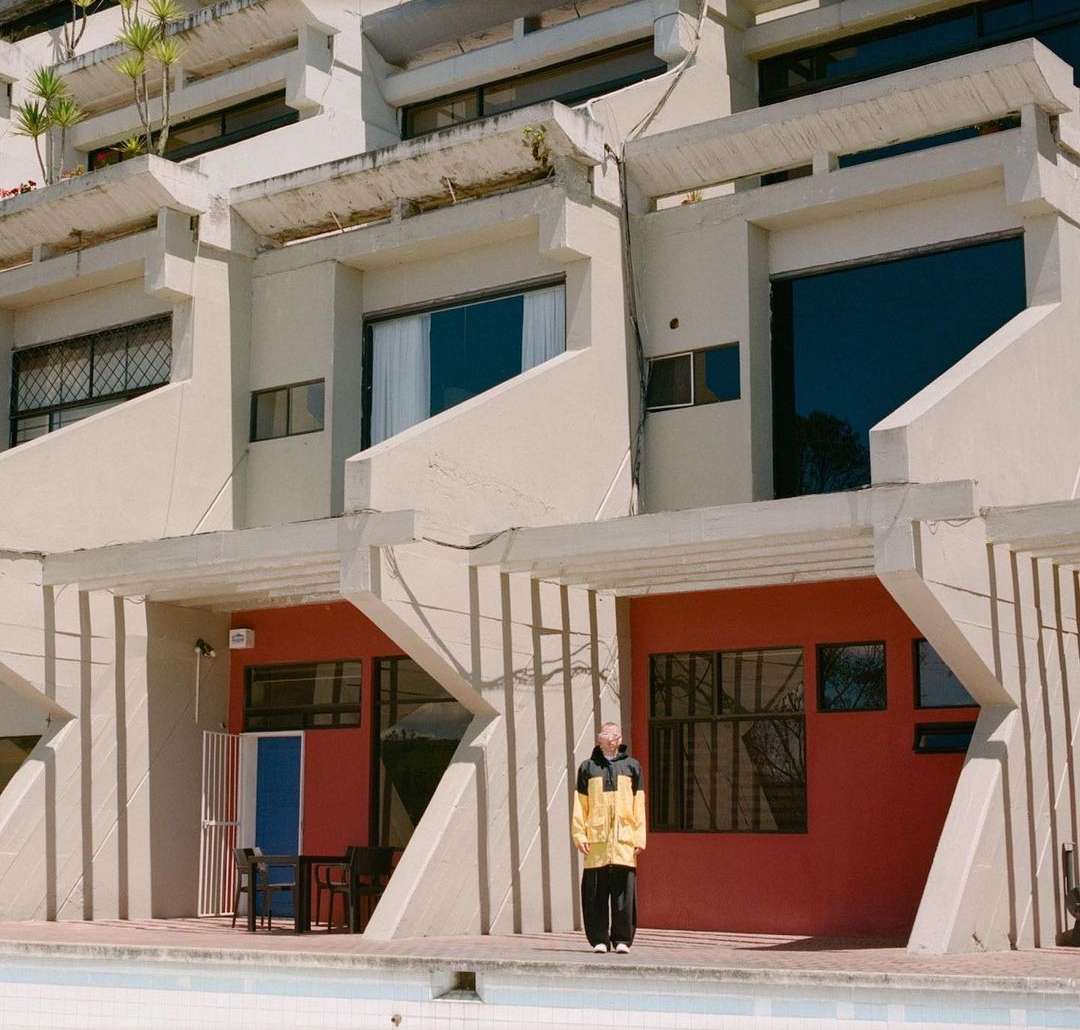 Person in yellow raincoat stands near a pool in front of a modern architectural building on a sunny day, capturing an urban scene.