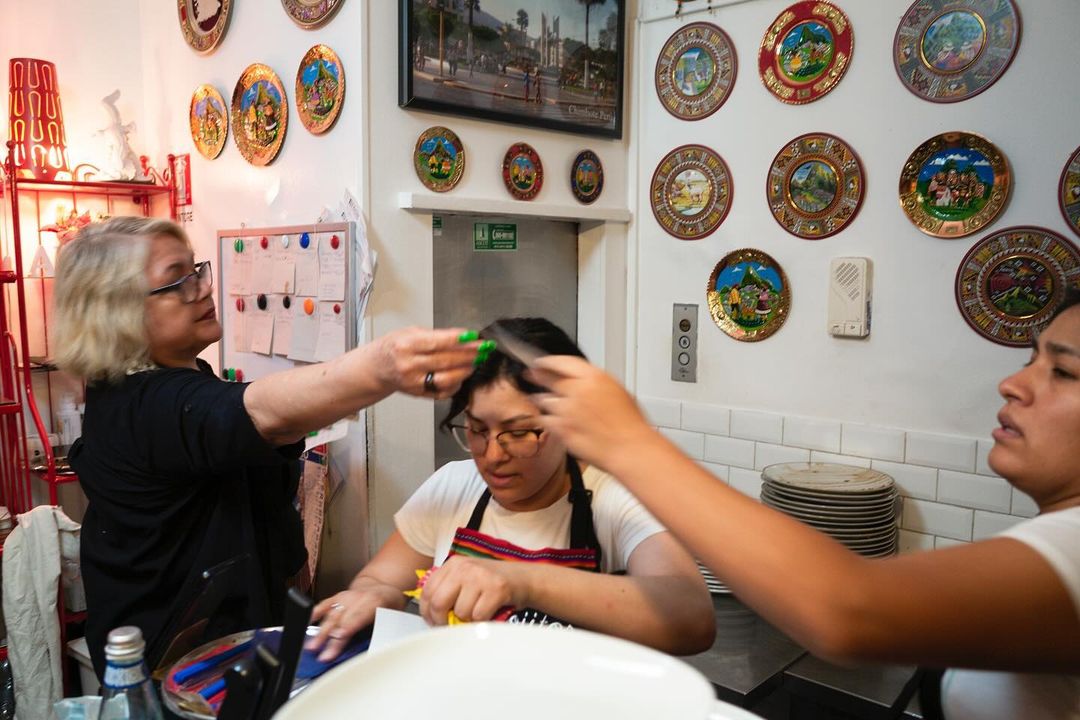 Three women work in a kitchen: one combs another woman's hair while the third woman looks on, surrounded by plates on the wall.