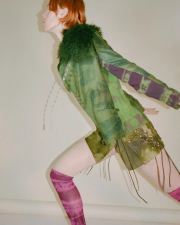 A slender red-haired model poses in a green jacket with a furry collar, matching shorts, patterned pink socks, and white tights.