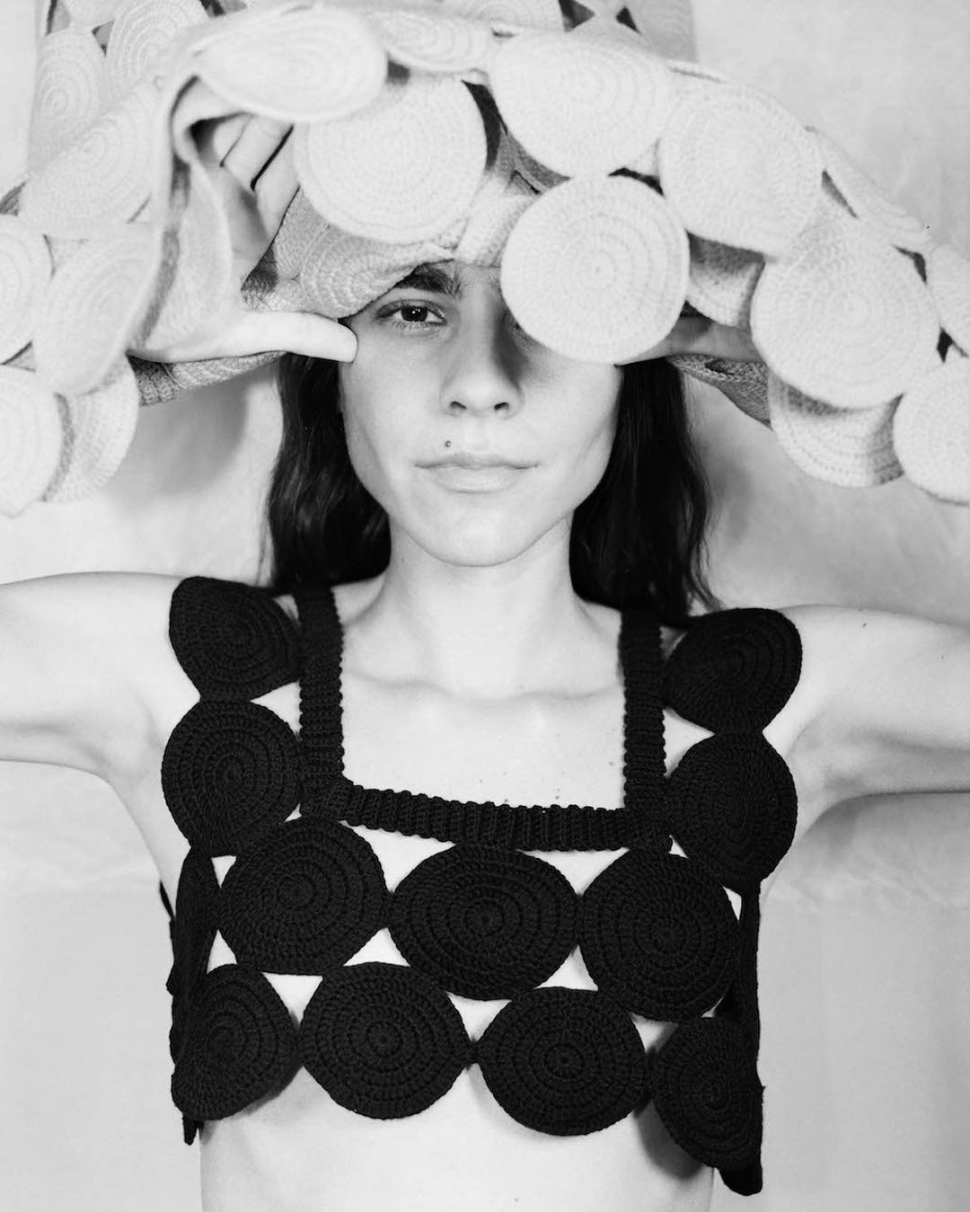 Black and white portrait of a woman wearing a crocheted top and a hat with circular embellishments, in a neutral indoor setting.