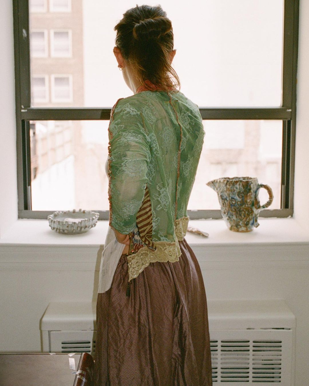 Back view of a woman in a green lace top and dark skirt standing by a window in an interior setting, looking out towards city buildings.