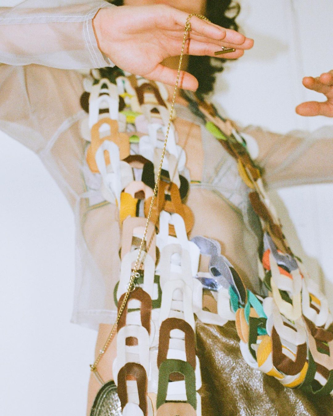 A person wearing an oversized necklace poses in front of a white wall with an indistinct expression and a gold chain.