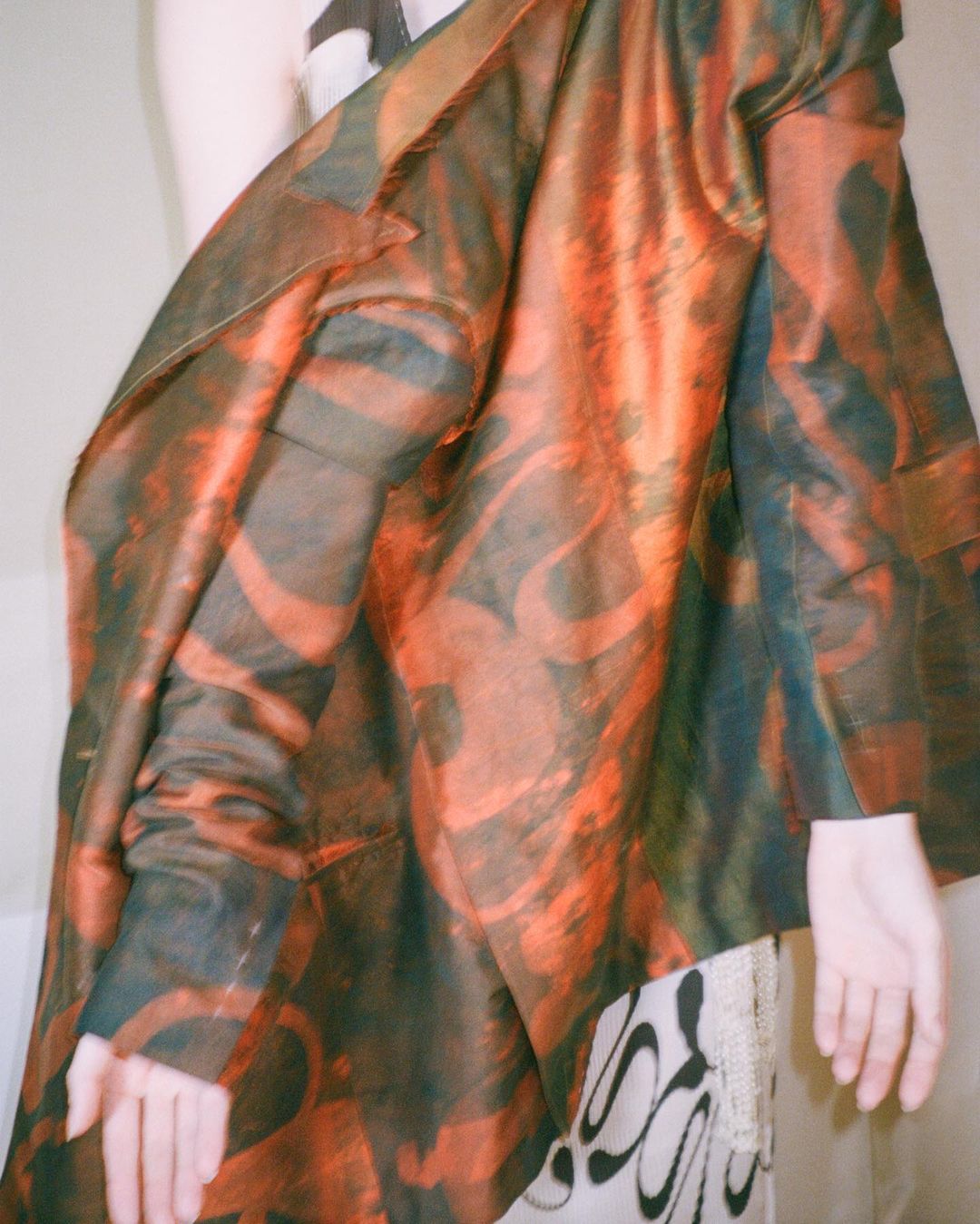 Person partially visible wearing a printed blazer with swirling patterns in orange and dark green, posing indoors in soft lighting.