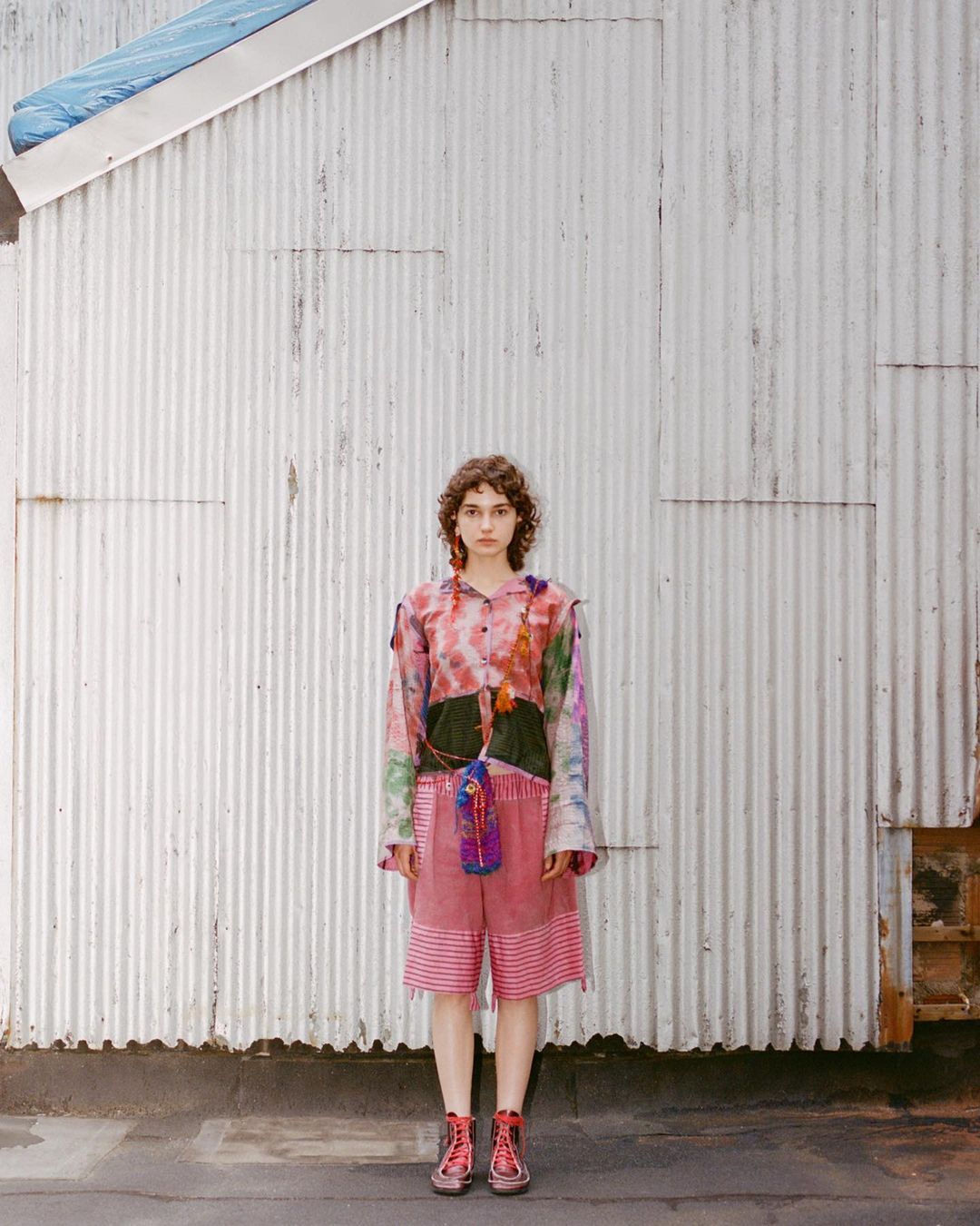Adult female stands outside on concrete wearing an artsy tie-dye shirt, red shorts, and red boots with a corrugated metal wall backdrop.