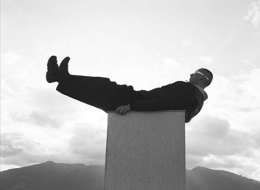 Man in sunglasses and suit lies on a concrete pillar with his legs in the air, monochrome shot against a cloudy sky and distant mountains.