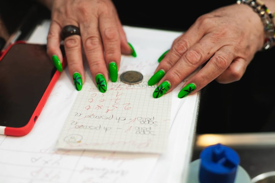 Adult hands with bright green tropical nail art place coin on paper; red smartphone and blue object in soft-focus background.