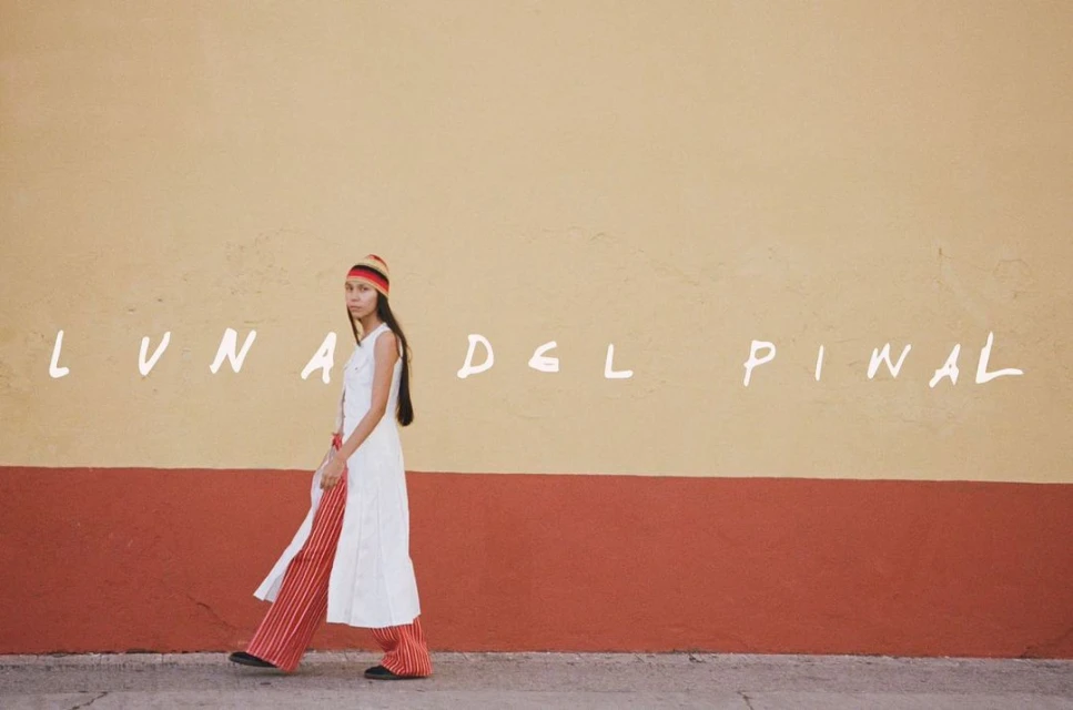 Woman walks in front of a colorful yellow and red wall wearing a white dress, red striped pants, and a knit cap in a fashion shot.