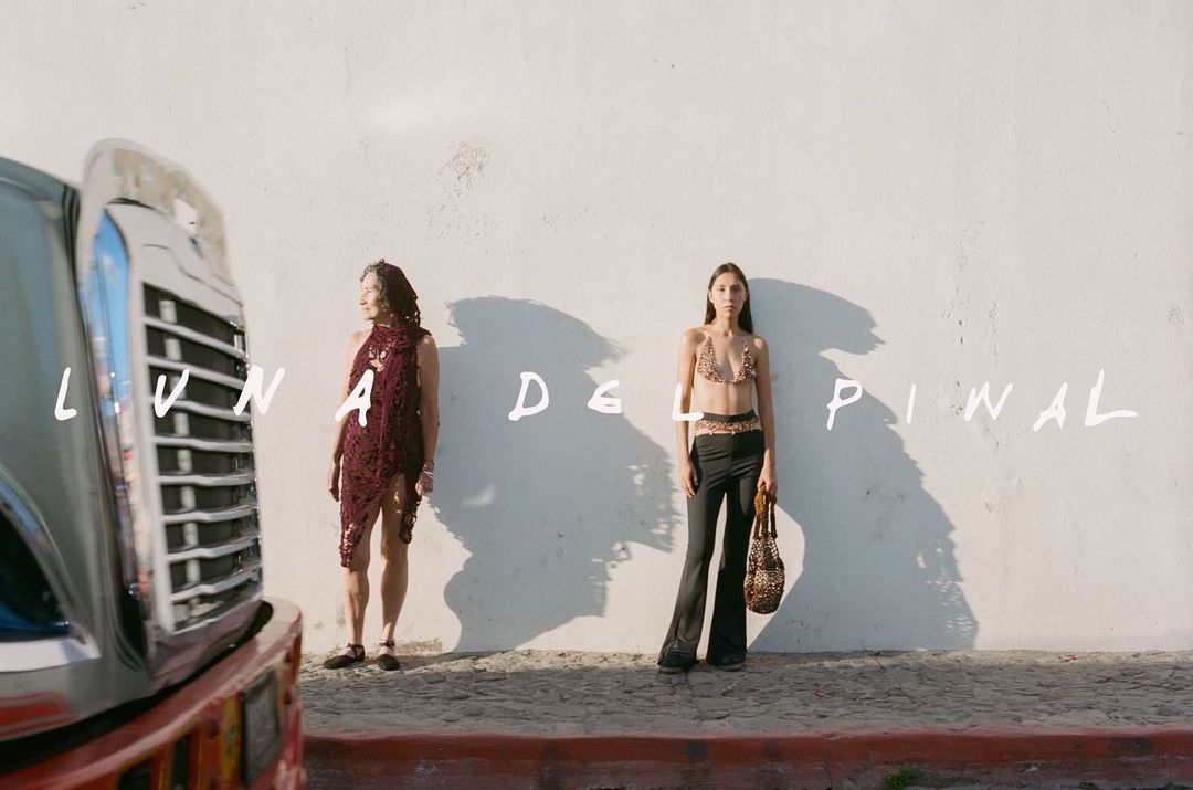 Two women stand against a weathered wall, one in a maroon crochet dress, the other in a crochet top and pants, with a classic car.