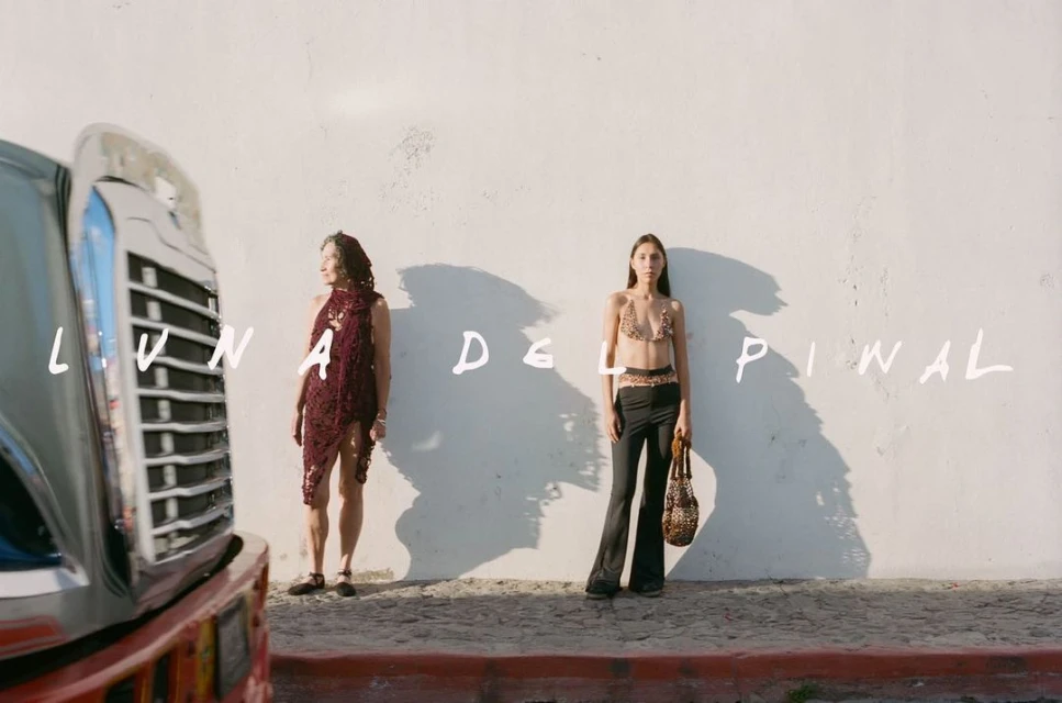 Two women stand against a weathered wall, one in a maroon crochet dress, the other in a crochet top and pants, with a classic car.