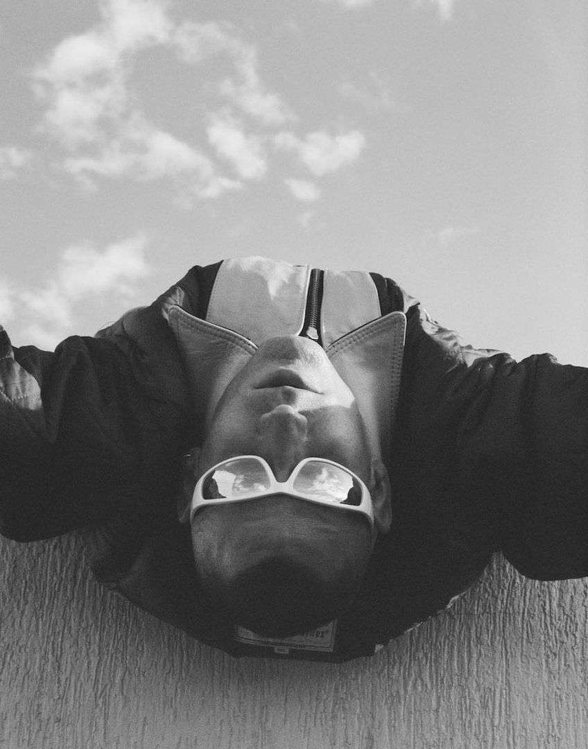 Black and white portrait of a man wearing sunglasses and jacket against a sky with clouds, taken from a low angle.