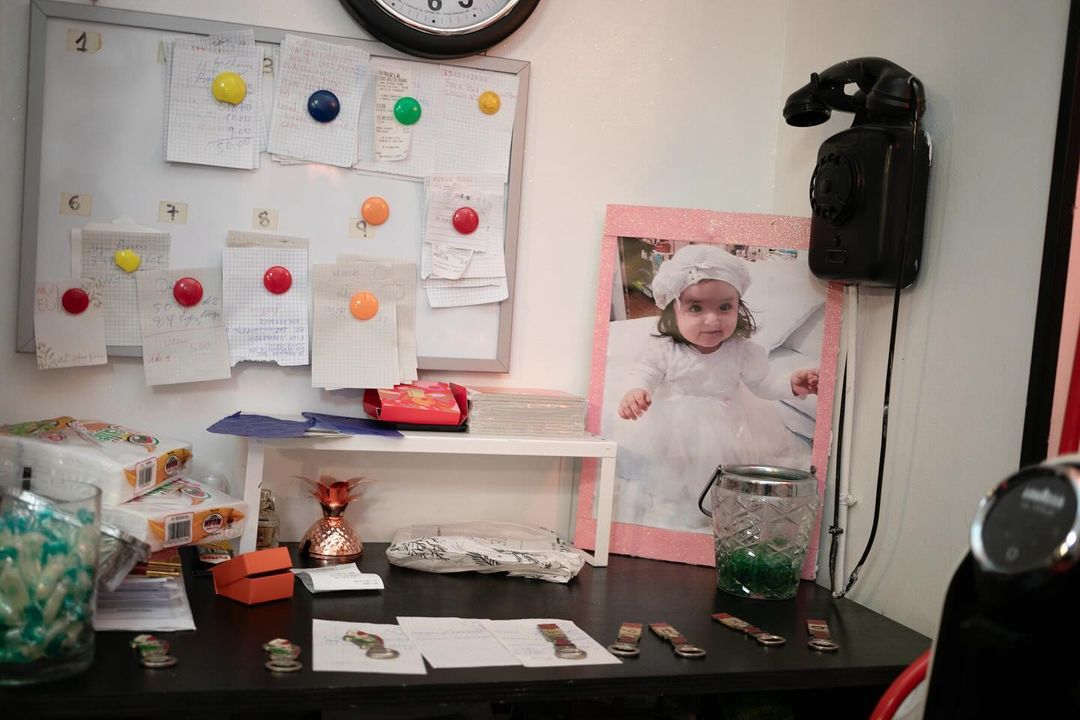 Indoor eye-level shot captures a photograph of a baby girl in a white dress with surrounding decorations, office supplies, and accessories.