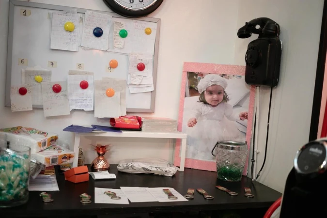 Indoor eye-level shot captures a photograph of a baby girl in a white dress with surrounding decorations, office supplies, and accessories.