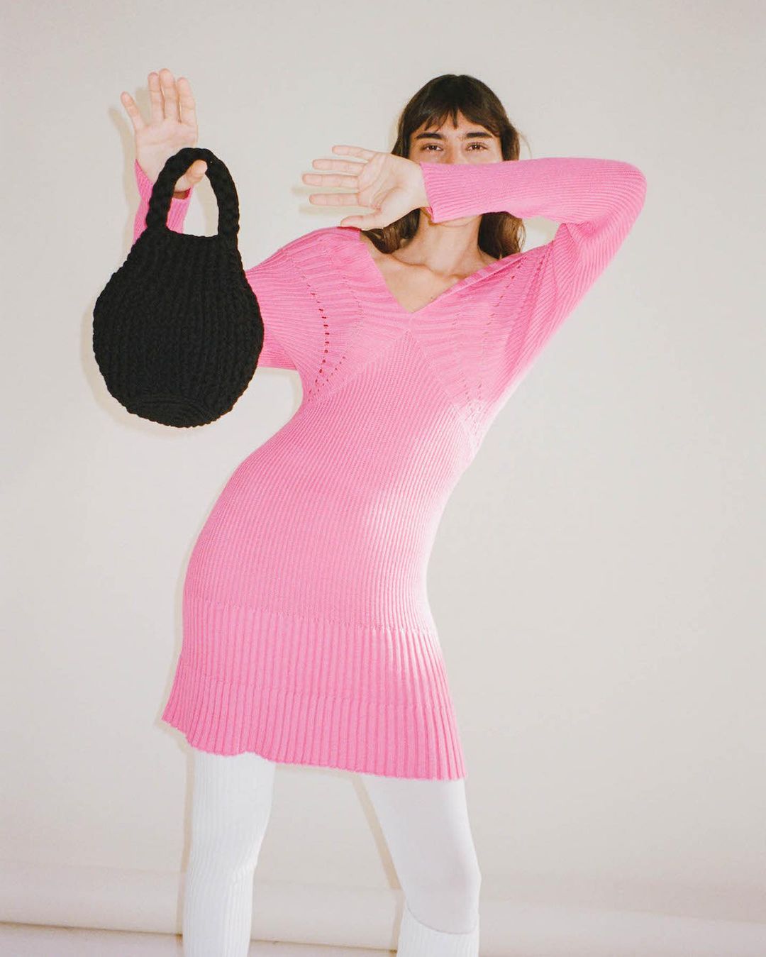 A woman in a pink dress poses with a black handbag in a studio, her hand raised towards her face in a light-hearted gesture.