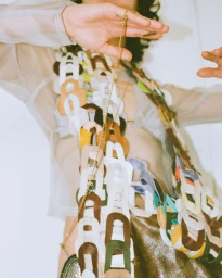 Model wearing a sheer top and holding a delicate gold chain necklace with a colorful leather link strap