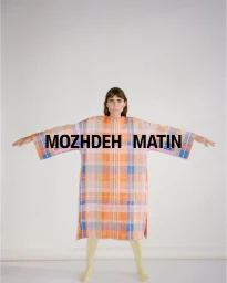 Model standing with arms outstretched wearing a plaid tunic against a plain white studio backdrop.