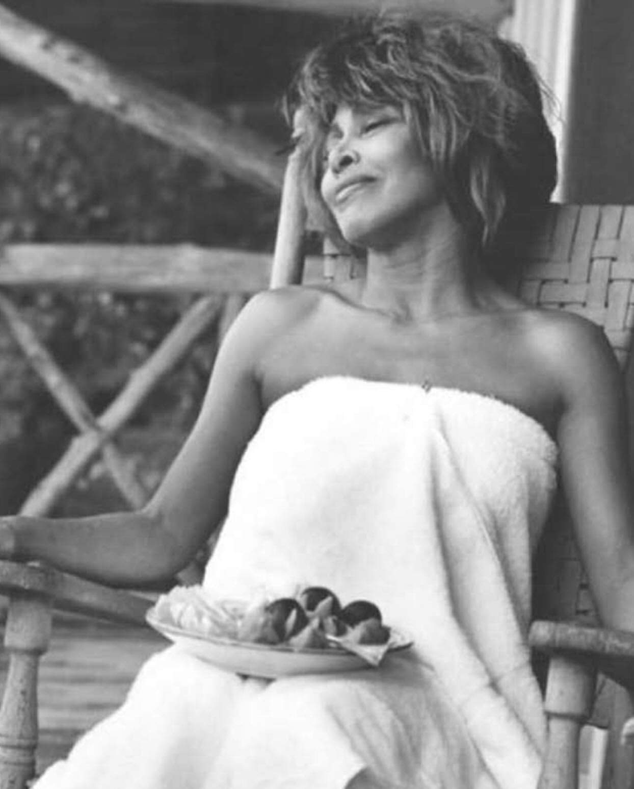 Black and white portrait of Tina Turner sitting in a wicker chair wrapped in a white towel and holding a plate of food.