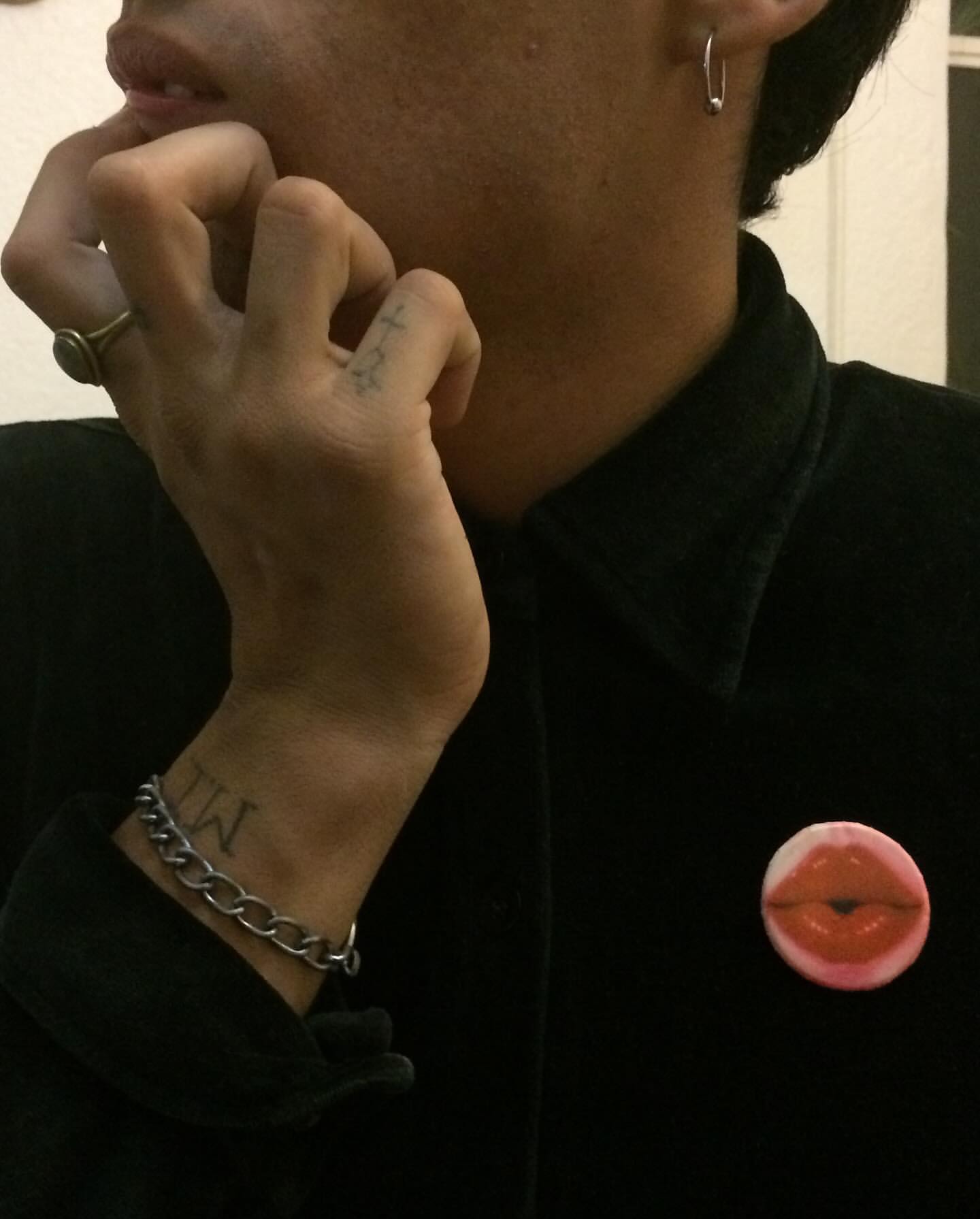 A person is photographed indoors, wearing a black shirt and silver chain bracelet, with an earring and ring visible, posing.