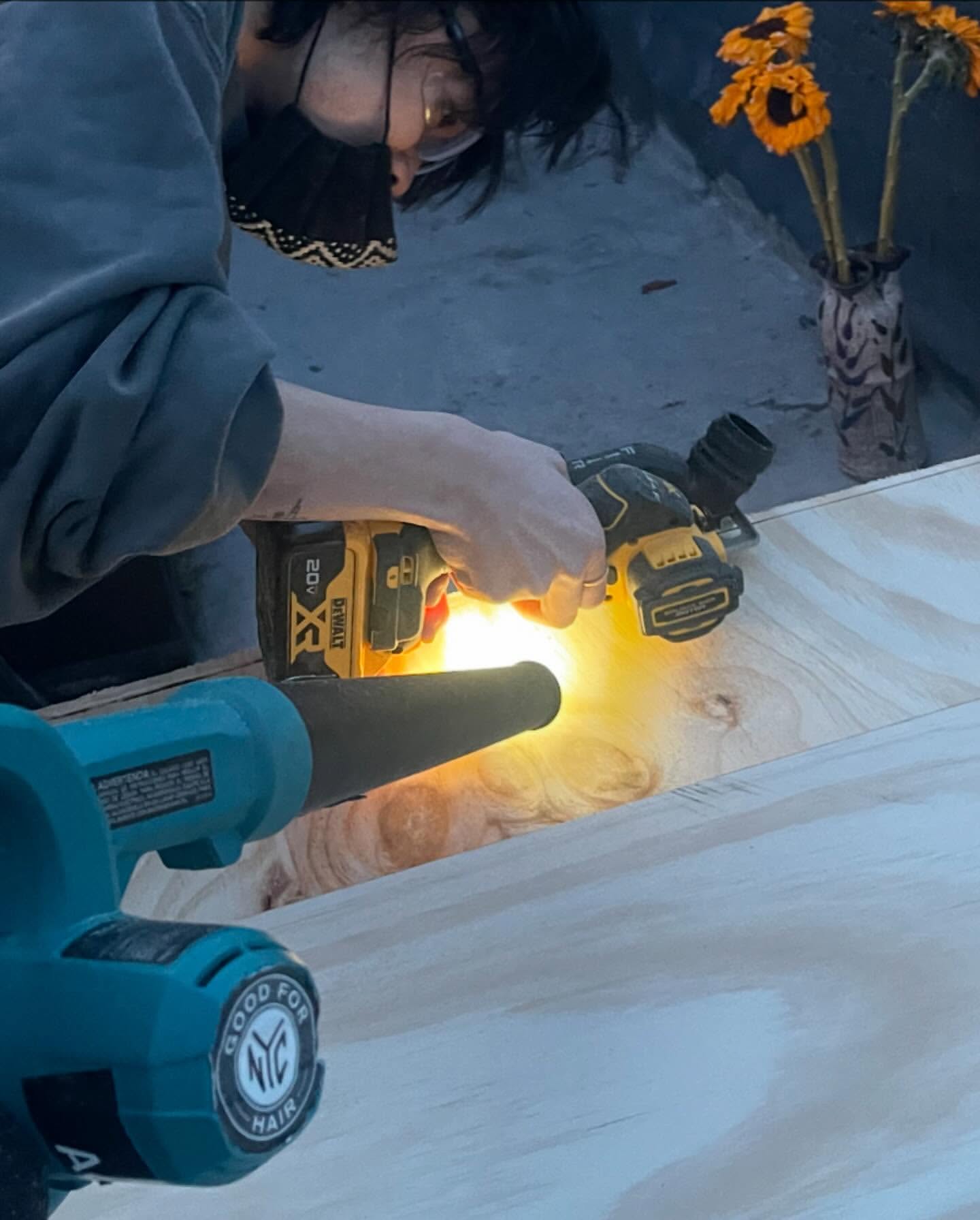Person wearing a mask and glasses is cutting wood with a circular saw near another tool and a vase of sunflowers indoors.