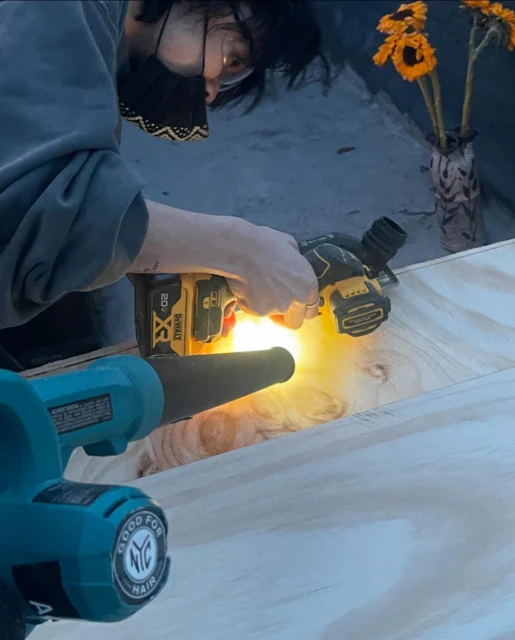 Person wearing a mask and glasses is cutting wood with a circular saw near another tool and a vase of sunflowers indoors.