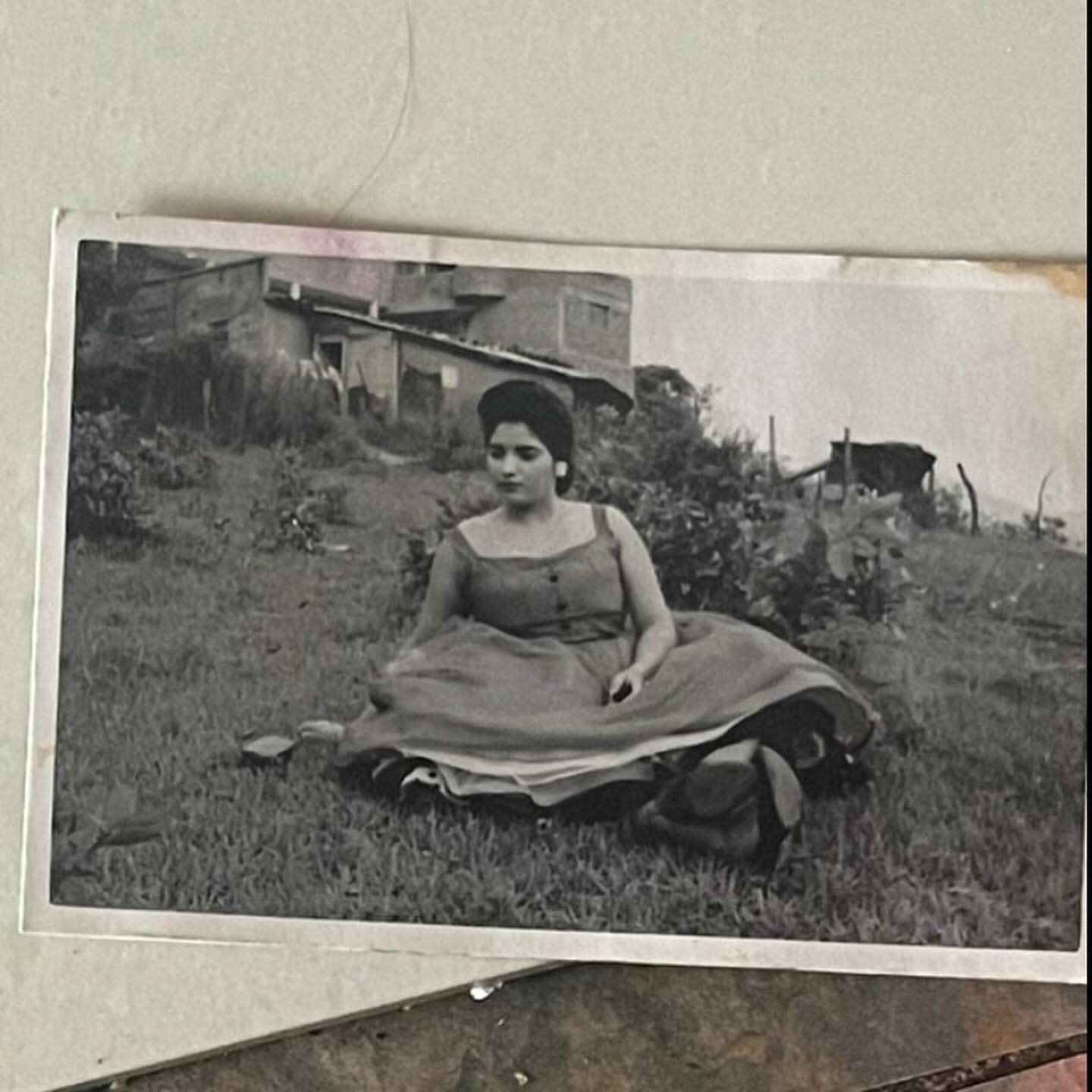 Vintage black and white photograph of a woman sitting on a grassy hill in a sundress with a building in the background.