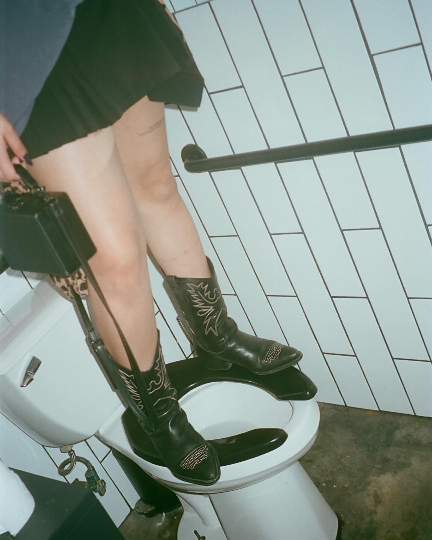 Unconventional fashion: Woman in a skirt and cowboy boots perches on a toilet, holding a purse in a bathroom with tiled walls.