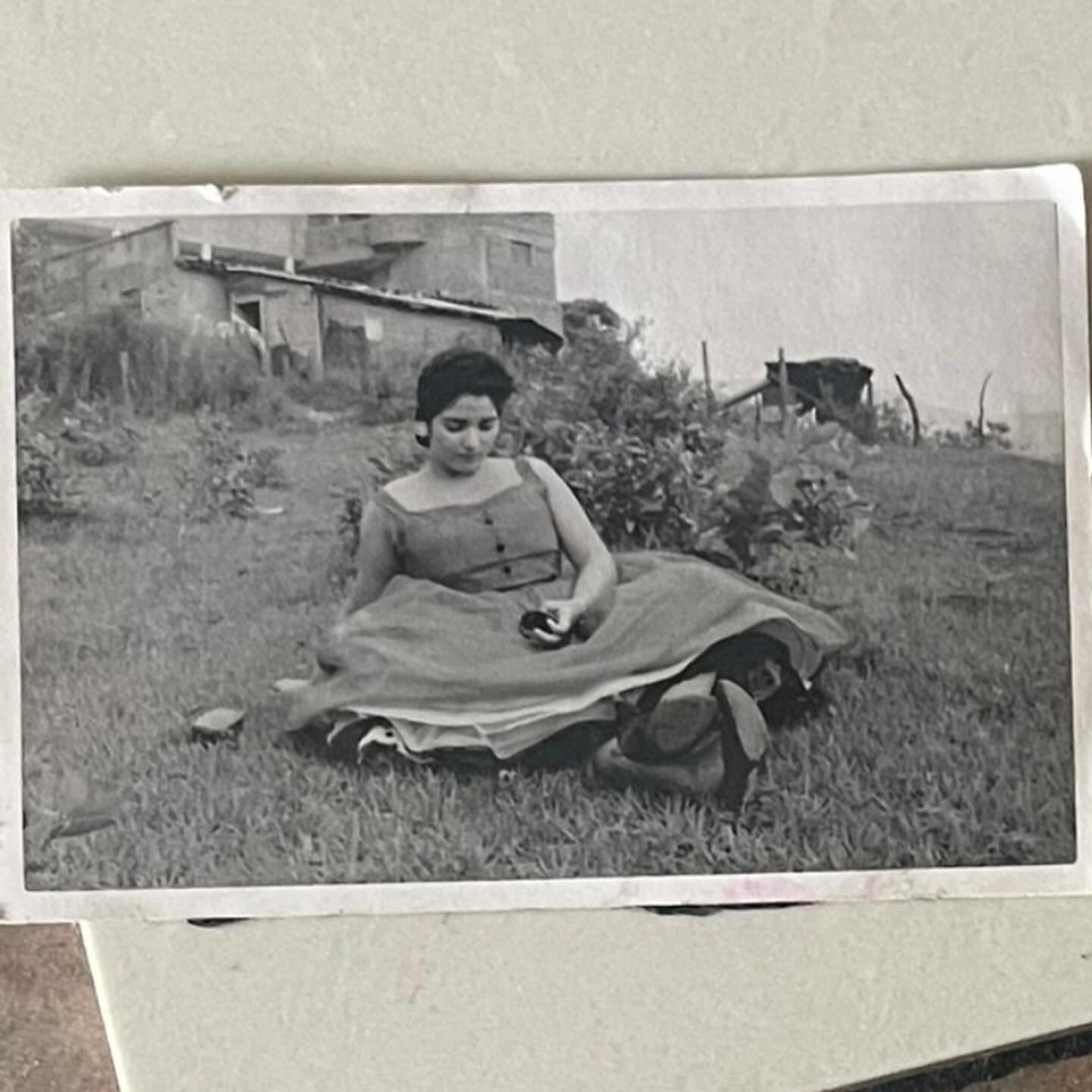 Vintage photo of a young woman in a dress sitting in a grassy field with a building and vegetation in the background.