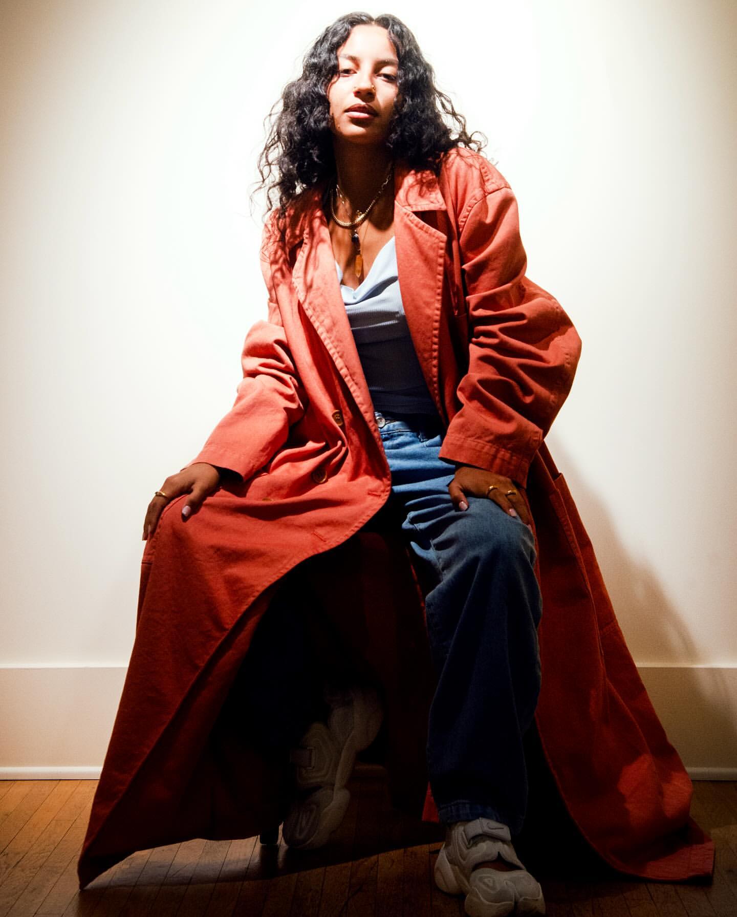 A woman with curly hair is sitting and wearing a long red coat and blue jeans, set against a neutral wall and wooden floor.