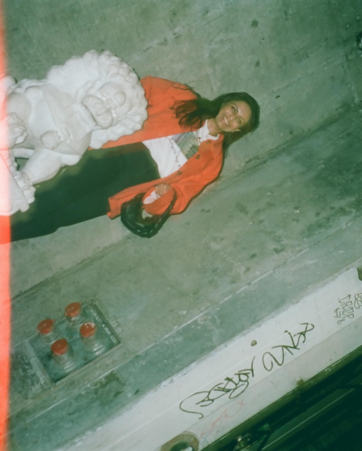 Young woman smiles in a concrete indoor space, lying near a white lion statue, wearing an orange jacket, capturing an authentic, candid moment.